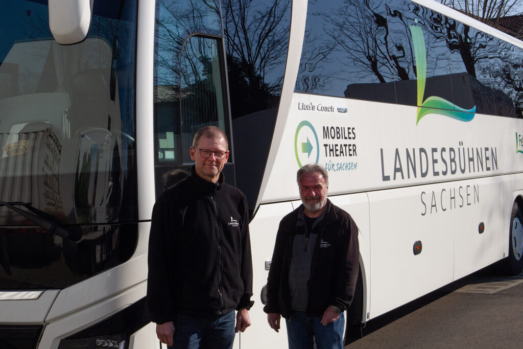 Zwei Männer stehen vor einem weißen Reisebus der Landesbühnen Sachsen.