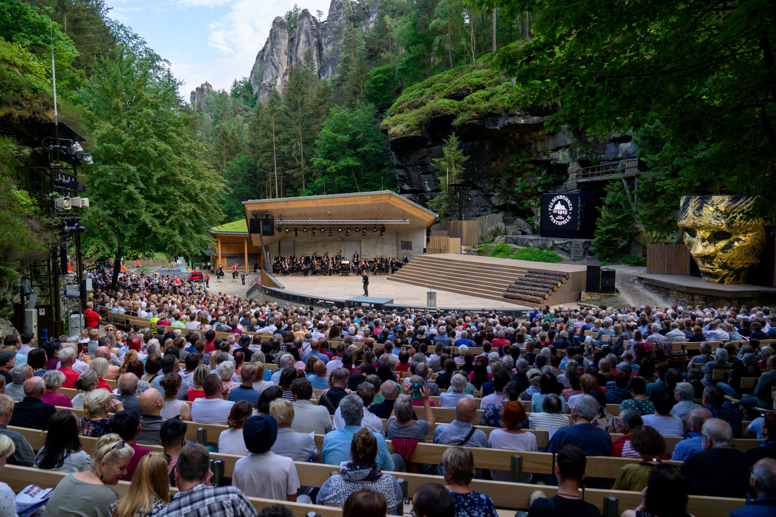 Ein Blick aus dem komplett gefüllten Zuschauerraum der neu gebauten Felsenbühne Rathen auf die Bühne mit Moderator und Orchester, während der Eröffnungsgala - Link zu Felsenbühne Rathen