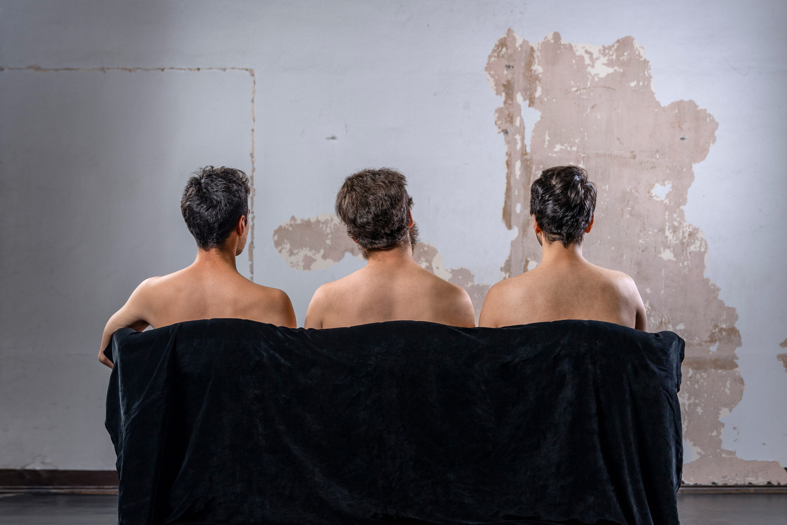 Drei Männer mit nacktem Oberkörper sitzen mit den Rücken zum Betrachter auf einem Sofa und gucken auf eine renovierungsbedürftige Wand.