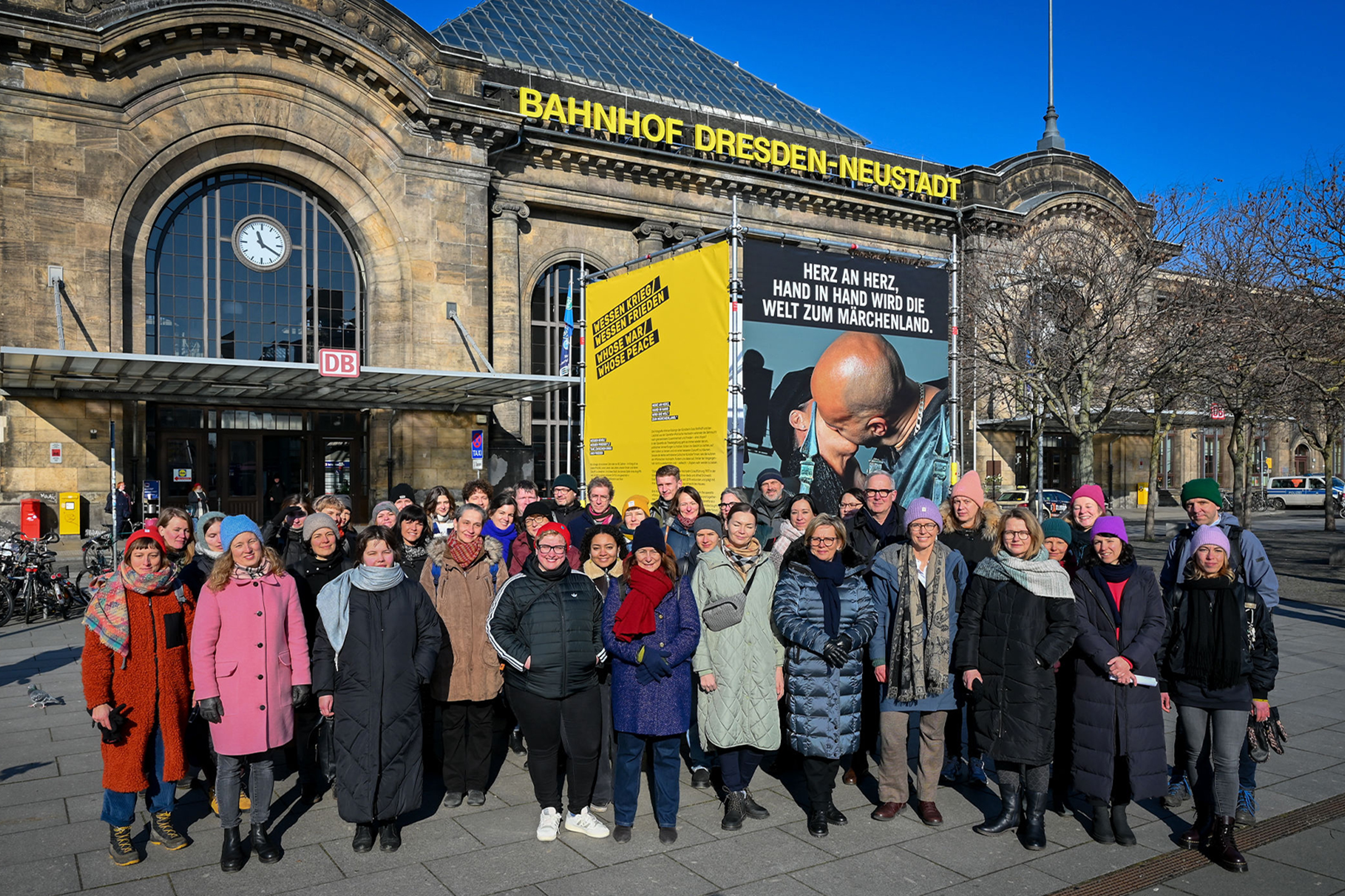 Eine große Personengruppe steht vor dem Bahnhofsgebäude Dresden-Neustadt. Auf dem Platz ist auch eine Plakataktion zum Thema "Weltoffenes Dresden" zu sehen.